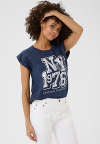 Tumši zili T-krekls ar sarullētām piedurknēm, uz kura redzams balts grafisks teksts "NY 1976 NACIONĀLĀ ČEMPIONĀTĀ". Apvienots ar augsto jostasvietu baltām džinsām.