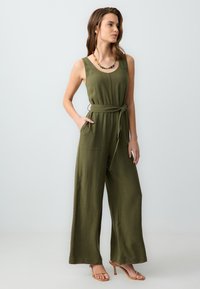 Combinaison vert olive à jambes larges, sans manches, avec poches avant et une ceinture nouée à la taille. Comprend un collier en perles multicolores.