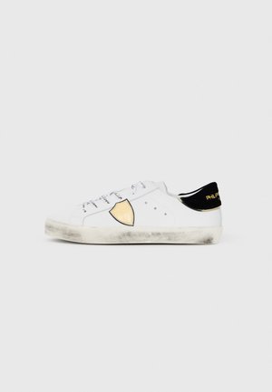 Witte sneaker met een gouden schildontwerp, zwarte hakaccent, gestructureerde rubberen zool en vetersluiting met merkveters. Iets gedragen.