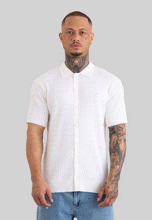 Mann mit Tattoos trägt weißes, strukturiertes Kurzarmhemd mit Kragen und Knöpfen, kombiniert mit hellblauen Jeans, steht vor grauem Hintergrund.