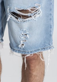 Svijetloplave traper kratke hlače s višestrukim izrezima i velikim rupama. Rubovi su neuređeni, s labavim koncem i ležernim krojem.