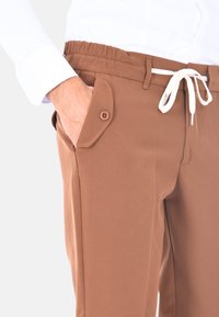 Pantaloni marroni con vita elastica e coulisse, dettagli con tasche laterali e texture liscia. Accentuati da un bottone per una chiusura sicura.