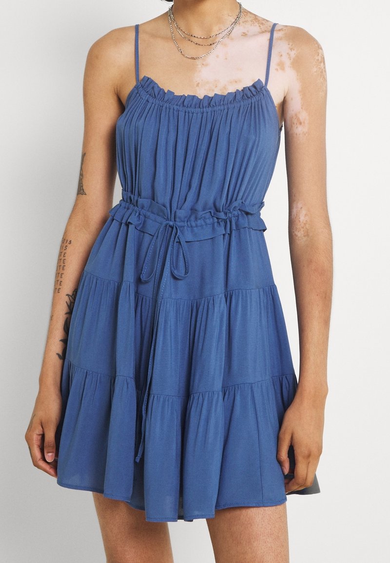 Personne portant une robe bleue sans manches à volants avec des fines bretelles, une taille cintrée, une jupe à plusieurs couches, des tatouages visibles et du vitiligo sur les bras et l'épaule.