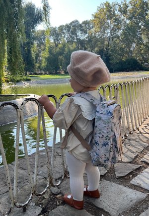 Bambino piccolo che indossa un basco beige e uno zaino floreale, mentre si tiene a una recinzione metallica bianca e guarda uno stagno circondato da alberi in un parco.