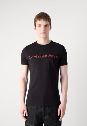 Unge mann iført en svart Calvin Klein Jeans T-skjorte med rød logo, stående foran en ensfarget hvit bakgrunn.