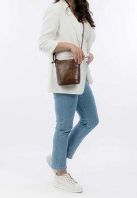 Donna che indossa un blazer bianco, jeans azzurri, sneakers bianche e porta una borsa a tracolla in pelle marrone dalla texture sulla sua parte sinistra.