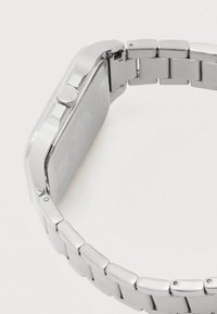 Sudraba metāla saite rokas pulkstenis skatīts no aizmugures ar plakanu korpusu un rievotu vainagu uz vienkārša balta fona.