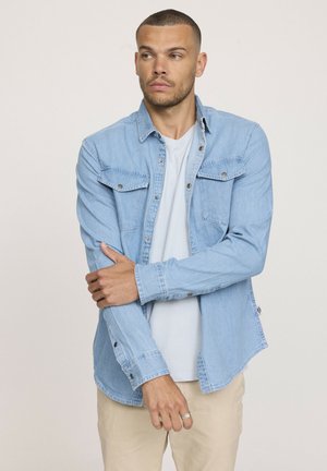 Chemise en denim bleu clair avec un devant à boutons, deux poches poitrine et des manches longues. Portée par-dessus un t-shirt de couleur claire.