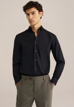 Schwarzes Langarmshirt aus glattem Stoff, mit klassischem Kragen und Knopfleiste, kombiniert mit grauen strukturierten Hosen.