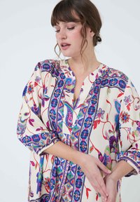 Bloemenpatroon tuniek in zachte crèmekleurige stof, met levendige paarse, roze en groene ontwerpen. V-hals en driekwartmouwen.