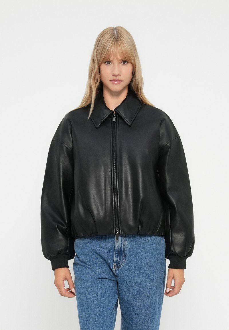 Schwarze Lederbomberjacke mit spitzem Kragen, Reißverschluss und übergroßen Ärmeln. Glatte Textur und gerippte Bündchen. Jeanshosen sichtbar.