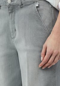 Szare dżinsy charakteryzują się luźnym krojem, wyraźnymi przednimi kieszeniami, zamkiem błyskawicznym oraz małą metką marki na prawej stronie pasa.