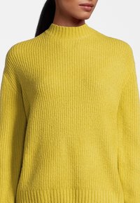 Gelbes Rippstrickpullover mit hohem Kragen, der eine lockere Passform und strukturierten Stoff aufweist. Der untere Saum ist leicht verlängert und hat Rippakzente.