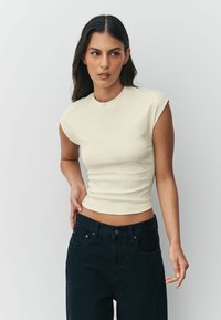Krém színű, testhez simuló póló sapkaszerű ujjakkal, kerek nyakkivágással és sima textúrával, magas derekú, sötét farmernadrággal párosítva.
