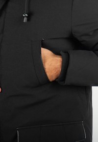 Doudoune noire avec une surface texturée, dotée d'une poche zippée au niveau de la poitrine et de poignets côtelés. Capuche ajustable avec cordon de serrage.