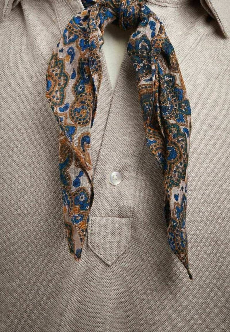 Beige textielhemd met een kleine kraag, voorzien van een patroon in bruin en blauw dat rond de hals is geknoopt. Inclusief een centrale knop en zak.