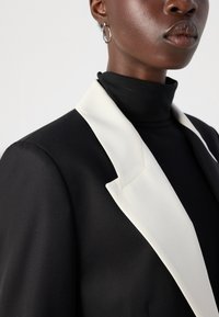 Black blazer with a white shawl collar, featuring smooth fabric, structured shape, and close-fitting high neck. Silver hoop earring visible.