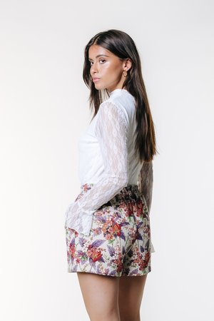 Biely top s dlhým rukávom z čipky s vysokým golierom, skombinovaný s kvetovanými šortkami s ružovými, fialovými a zelenými vzormi na svetlom pozadí.