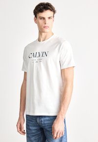 Camiseta blanca de algodón con el logo "CALVIN" en azul marino, cuello redondo y mangas cortas. Combinada con jeans de mezclilla azules.