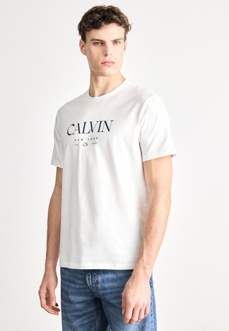 Camiseta blanca de algodón con el logo "CALVIN" en azul marino, cuello redondo y mangas cortas. Combinada con jeans de mezclilla azules.