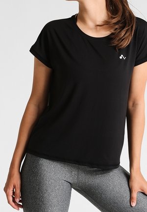 Femme portant un haut noir à manches courtes et des leggings gris, avec les cheveux attachés en queue de cheval basse, debout devant un fond uni.