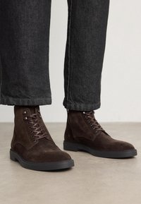 Bottines en daim marron à bout rond, design à lacets, semelle en caoutchouc, associées à un jean denim foncé, mettant en valeur un ourlet brut et une coupe ajustée.