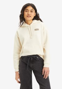 Krämmfärgad hoodie med dragsnören, känguru ficka och en brun Levi's-logga. Kombinerad med högmidjade svarta jeans. Slät textur.