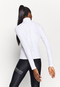 Biała, dopasowana bluzka z długim rękawem i golfem, wykonana z gładkiej tkaniny. Do kompletu czarne legginsy z nadrukowanym logo po boku.