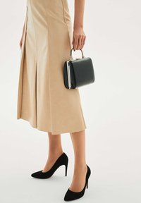 Czarna skórzana torebka z srebrną klamrą, trzymana w dłoni. Osoba ma na sobie beżową spódnicę do kolan oraz czarne szpilki.