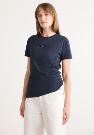 T-shirt in cotone blu navy con logo "BOSS" in paillettes. Maniche corte e scollatura rotonda. Abbinato a pantaloni di colore chiaro.