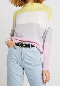 Pull en tricot à rayures jaunes, grises et roses, col montant et coupe ample. Associé à un jean bleu clair et une ceinture noire avec une boucle dorée.