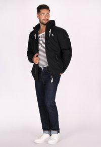 Giacca nera con cappuccio, camicia a righe, jeans scuri e sneakers bianche; presenta cerniere, cordini e dettagli con logo sulla giacca.