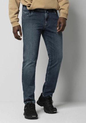 Homme portant un jean bleu délavé, un sweat à capuche beige et des baskets Balenciaga noires, debout devant un fond gris clair uni.