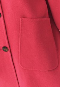 Manteau en laine rose vif avec une poche proéminente, des bords arrondis et une fermeture à boutons. Texture douce avec une apparence nette et ajustée.