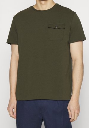 Homme portant un t-shirt à manches courtes vert olive avec poche poitrine à boutons-pression et un pantalon bleu marine, debout devant un fond uni.