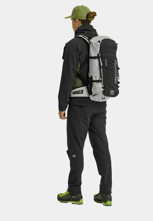 Back view of a person wearing a black and gray Ortovox backpack, designed with adjustable straps, patterned fabric, and green accents.