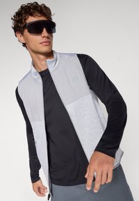 Gray and black athletic vest with zip front, lightweight fabric, textured panels, and long-sleeved black base layer. Dark sunglasses worn.