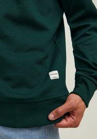 Uomo in felpa verde scuro con un piccolo logo bianco di Jack & Jones vicino all'orlo, che tiene il bordo della felpa con la mano destra.