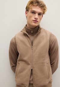 Beige Weste mit Reißverschluss und hohem Kragen, aus weichem Stoff. Verfügt über Seitentaschen und ein minimalistisches Design. Wird über einem passenden Langarmoberteil getragen.