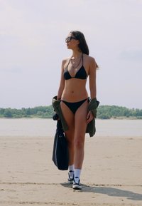 Čierny bikini set, svetlohnedá bunda prehodená cez rameno, tenisky s bielymi ponožkami a elegantné slnečné okuliare. Plážové prostredie s vodou v pozadí.