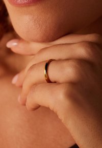 Goldring mit einer glatten, polierten Oberfläche, getragen an einem Finger; weicher, natürlicher Hautton und hellrosane Nägel im Hintergrund.