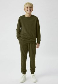 Olivgrön fleece-tröja med långa ärmar och matchande joggerbyxor; båda har elastiska manschetter och avslappnad passform, tillsammans med vita sneakers.