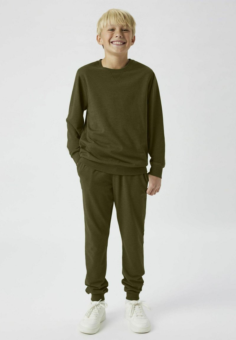 Olivgrön fleece-tröja med långa ärmar och matchande joggerbyxor; båda har elastiska manschetter och avslappnad passform, tillsammans med vita sneakers.