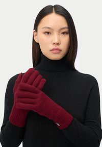 Burgundy stickade handskar med ribbade ärmar, mjuk textur och en logotypetikett, bärs av en modell i en svart polotröja.