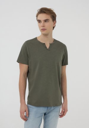 T-shirt verde oliva a maniche corte con scollatura a henley e due bottoni. Tessuto morbido, vestibilità rilassata, abbinato a jeans azzurri chiari.