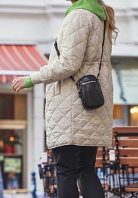 Giacca beige trapuntata con una felpa verde sotto, borsa a tracolla nera con zip dorata e pantaloni scuri. Design caldo e casual.