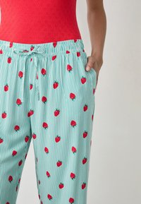 Pantaloni leggeri a righe teali con motivi di fragole rosse. Presentano una cintura elastica con cordoncino regolabile e tasche laterali.