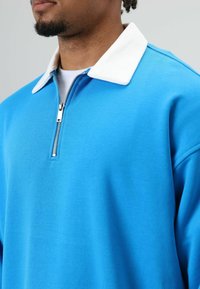 Blauwe pullover met een witte kraag, voorzien van een halve rits bij de hals. Gemaakt van zachte stof met een ontspannen pasvorm en een gladde textuur.