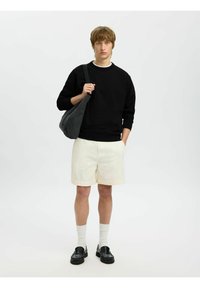 Sudadera negra, pantalones cortos color crema, bolso gris, mocasines negros y calcetines blancos. El modelo se encuentra sobre un fondo liso, con las manos en los bolsillos o sosteniendo el bolso.
