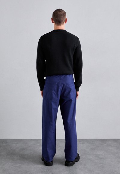 Svart stickad tröja, blå vida byxor med synliga sömnadsdetaljer och kraftiga svarta skor. Bakifrån mot en neutral bakgrund.